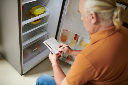 Aged Man Writing Shopping List For Grocery Store When Checking Fridge In Kitchen