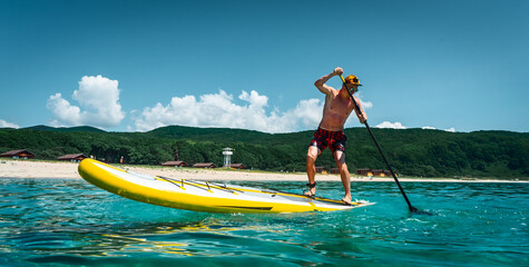 Tanned man stands on a yellow supboard and paddles. Active sport on the beach in the turquoise sea water - obrazy, fototapety, plakaty