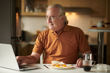 Smiling elderly man eating breakfast and checking e-mails on laptop