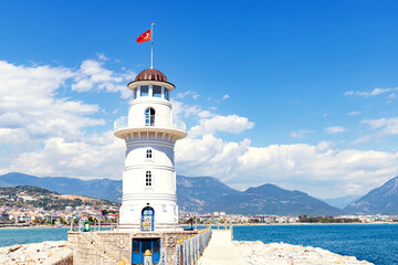 Fototapeta na wymiar White lighthouse with turkish flag in Alanya, Turkey. Panoramic view of city, sea and harbour under cloudy sky. Selective focus.