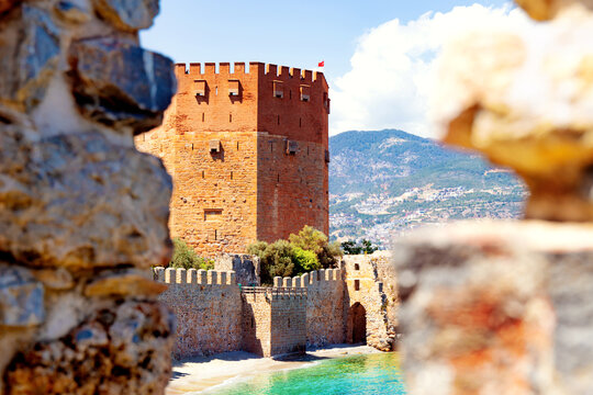 Beautiful view of ancient Red Tower in Alanya, Turkey, between two merlons of old stone wall. Sandy beach under old historic walls on sunny day. Blurred foreground. Selective focus.