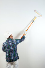 A male builder in a hard hat paints a white wall with a roller