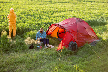 Young couple cooking food, spending summer time together at campsite on a green field. Man and...