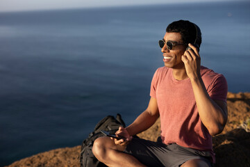 Young man sitting on the stone listening the music. Tourist man on the top of the mountain