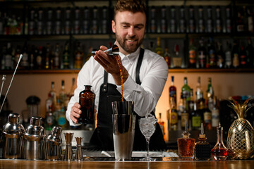 various shakers and bottles stand on the bar counter and male bartender masterfully pours alcoholic drink into cup
