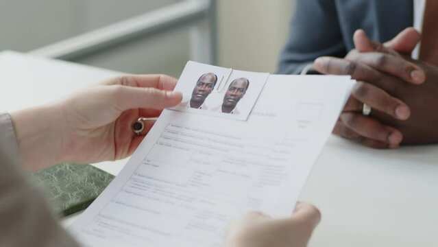 African American man in formalwear giving documents, photos and application form to female visa officer while having interview in embassy