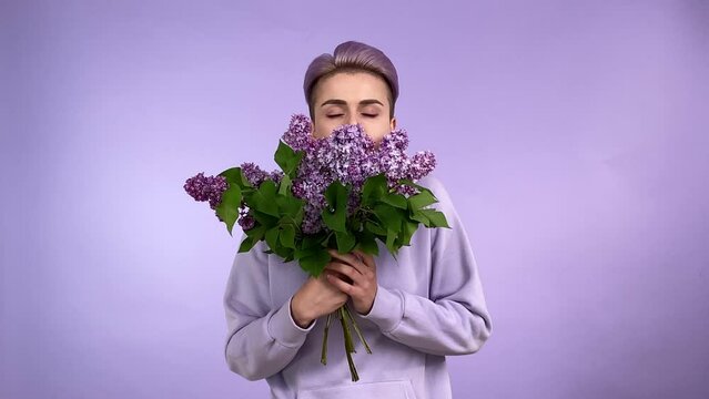 Charming young adult woman with trendy short dyed violet hair, holding smelling lilac bouquet, enjoying aroma flowers, wearing violet hoodie, isolated on purple background. Monochrome concept