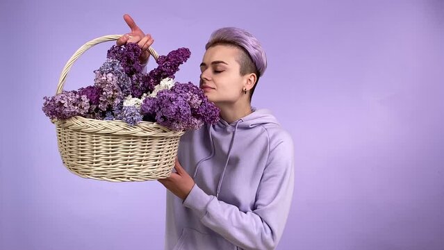 Cheerful young adult woman with trendy short dyed hair, holding wicker basket, smelling lilac bouquet, enjoying aroma flowers, wearing violet hoodie, isolated on purple background. Monochrome concept