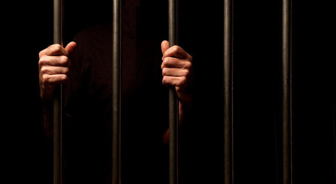 hands of a prisoner behind prison bars on black background
