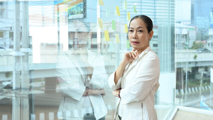 Attractive mature businesswoman taking break, resting and standing near large window at office with city buildings in background