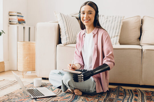Smiling cheerful young female student listening to online English lesson in her wireless earphones, noting down in copybook, having bionic cyber hand, sitting against white sofa, in front of laptop