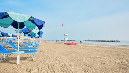Deckchair with colorful umbrellas on a sunny summer day. In the background a lifeboat next to a lifeguard tower on the shore of the Adriatic Sea in Italy.