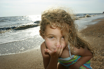 a little girl, standing on the seashore, sends an air kiss, wavy hair blowing in the wind. The...