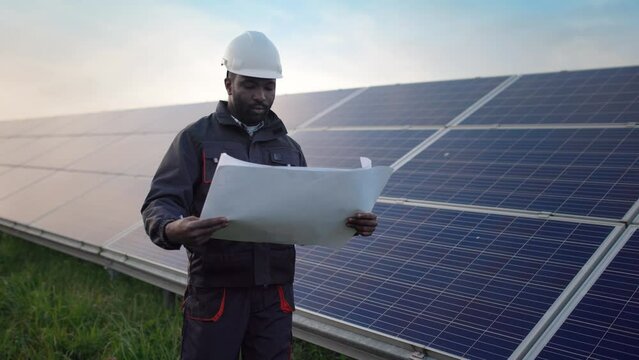 Young African-American engineer in protective helmet and uniform walks among the fields with solar panels with paper scheme in his hands studying location and serviceability of batteries.