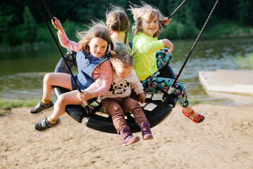 four children swinging on wicker swing on playground in park by lake, Sand and children's summer vacation with friends - obrazy, fototapety, plakaty