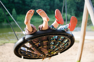 kids swinging on wicker swing on playground in park by lake, children's summer vacation with friends - obrazy, fototapety, plakaty