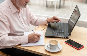 an unrecognizable older man sitting in a coffee shop or bar studying and using his laptop to make notes