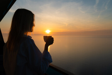 Happy young woman in a white shirt is drinking coffee from a white cup on the balcony, watching the...