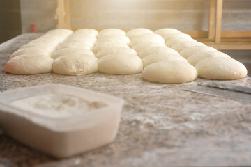 Fototapeta na wymiar On the table there is a lot of prepared dough for baking buns or other bakery products, a container with flour and a knife-cutter for dough. Photo with illumination