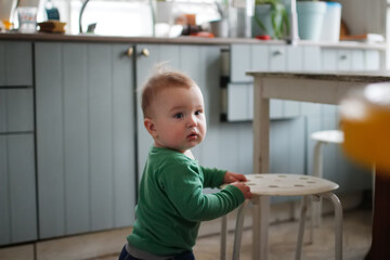 Funny one-year-old toddler standing at stool in kitchen, real people in real interior