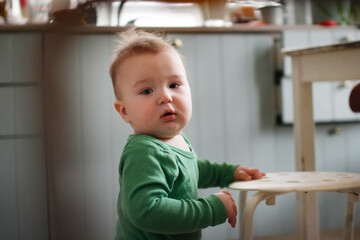 Funny one-year-old toddler standing at stool in kitchen, real people in real interior