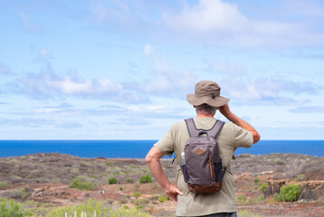 Fototapeta premium Rear view of active senior man with backpack and hat while hiking outdoors looking at the horizon. Elderly man enjoying retirement and healthy lifestyle. Blue sea and sky in the background