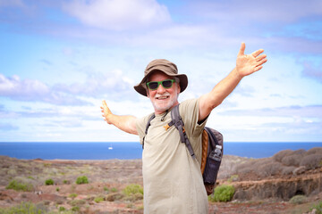 Happy active senior man with backpack and hat while hiking outdoors looking at camera with outstretched arms. Blue sea and sky in the background