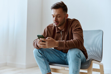 Friendly happy focused young man guy hold phone typing message chatting talking with family sitting on chair at home. Distance communication concept. Mobile App offer. Copy space