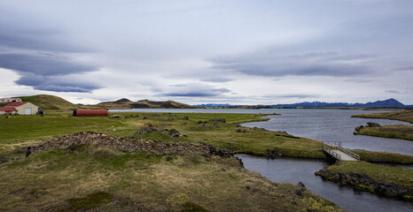Fototapeta na wymiar Farm buildings on the shore of a lake in Icelandic north. Volcanic crater and mountain range on the horizon.