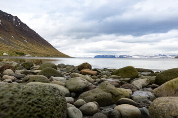 Fototapeta na wymiar Rocks and pebbles on the shore of Icelandic fjord. Slope of the mountain on the left. Mountain range on the horizon.