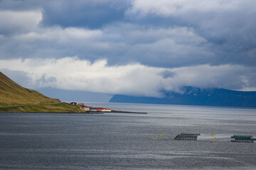 Industrial buildings on a slope of a cliff on Icelandic Atlantic shore. Fish farms close by. Volcanic mountains ranges in a distance.