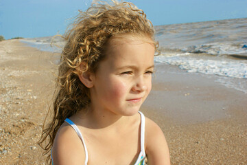 A little girl, thoughtful, sits on the seashore on the beach and looks into the distance. With flowing hair, portrait, postcard. The concept of vacation, travel, tourism, advertising, banner, mocap.