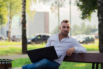 Thoughtful business man in a white shirt is working in the park with a laptop. Young man on the background of passing cars, hot sunny summer day. Warm soft light, close-up.