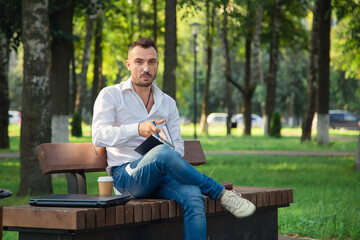 Obraz premium A serious man sits on a bench, makes plans, writes in a notebook. A young man on a background of green trees, a hot sunny summer day. Warm soft light, close-up.