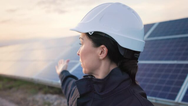 Close up of beautiful female caucasian engineer in protective helmet stands on background of solar panels and records video report looking at camera.
