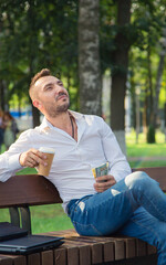 An agonizing man sits on a bench, drinks coffee, holds dollars. A young man on a background of green trees, a hot sunny summer day. Warm soft light, close-up.