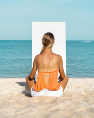 A blonde woman sits on a sandy beach in front of a white canvas.  The sea is in the background.