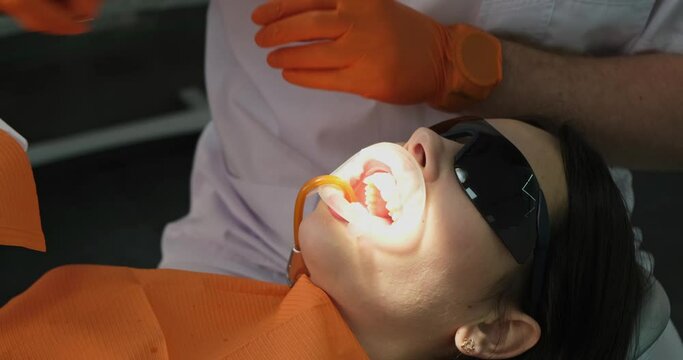 dental care, woman patient treats teeth at dentist close-up, female has saliva ejector and mouth expander in mouth, sunglasses are on her eyes from bright lighting of lamp in stomatologist office. 