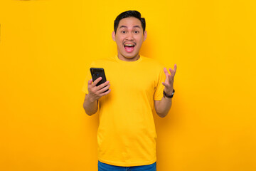 Shocked young Asian man in casual t-shirt holding mobile phone and looking at camera isolated on...