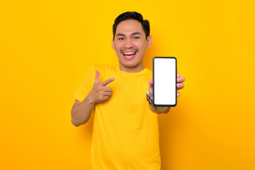 Mockup blank screen cellphone. Cheerful young Asian man in casual t-shirt showing mobile phone with blank screen, recommending mobile app isolated on yellow background. People lifestyle concept