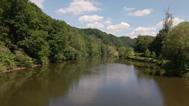 Lush, green nature surrounding a calm, brown river in North Rhine Westphalia, Germany on a sunny summer day. Ascending aerial fly over shot