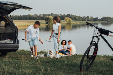 Two Teenage Children, Son and Daughter Playing with Pet Dog While Parents Having Picnic by the Lake, Four Members Happy Family Enjoying Weekend Road Trip by Car