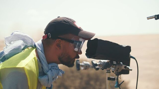 A cameraman in a reflective vest is watching the shooting in the desert. Film shooting.Trucks passing by