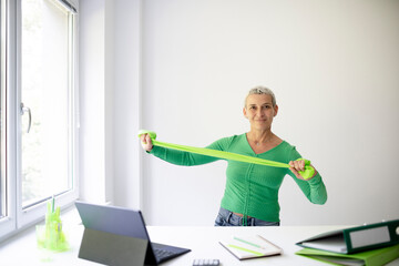 middle aged businesswoman standing at white high table in office doing stretching exercises with...