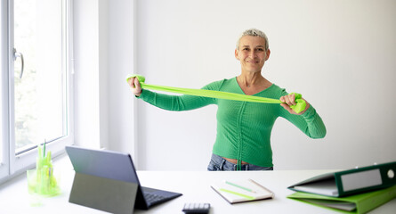 middle aged businesswoman standing at white high table in office doing stretching exercises with...