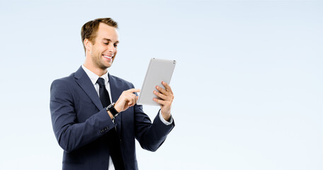 Happy smiling young business man in black suit, necktie using tablet pc computer, isolated over grey color background. Businessman holding touchpad at studio concept image.