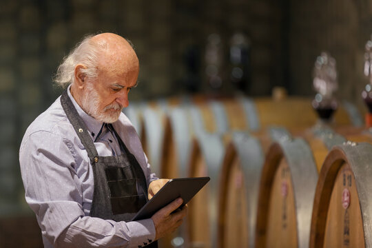 Caucasian experienced winemaker checking a wine aging in barrels data, using a touchpad tablet. Traditional and modern technology concept.