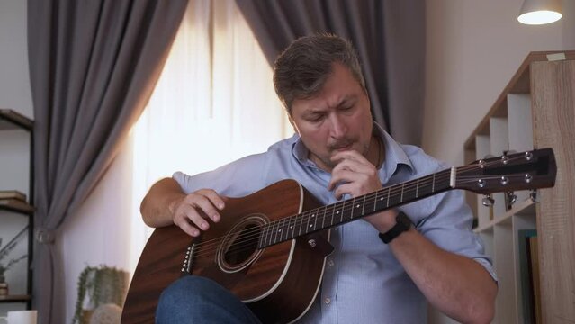 Musical hobby. Desperate man. Learning problem. Upset clueless middle-aged guy sitting sofa having trouble trying to play guitar in light room interior.