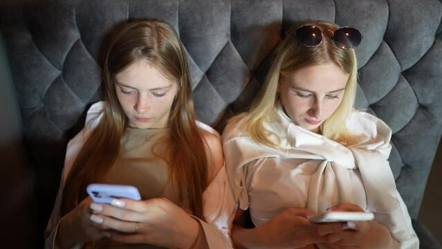 Relaxed two young girls using smartphone surfing social media, checking news, playing mobile games or texting messages sitting on sofa, close up