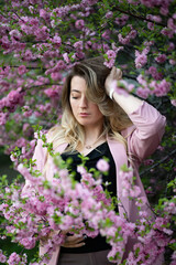 Fototapeta premium A very beautiful blonde in a pink jacket and a black blouse sensually poses standing among the flowers of cherry blossoms with her eyes closed. Goddess of nature or earth